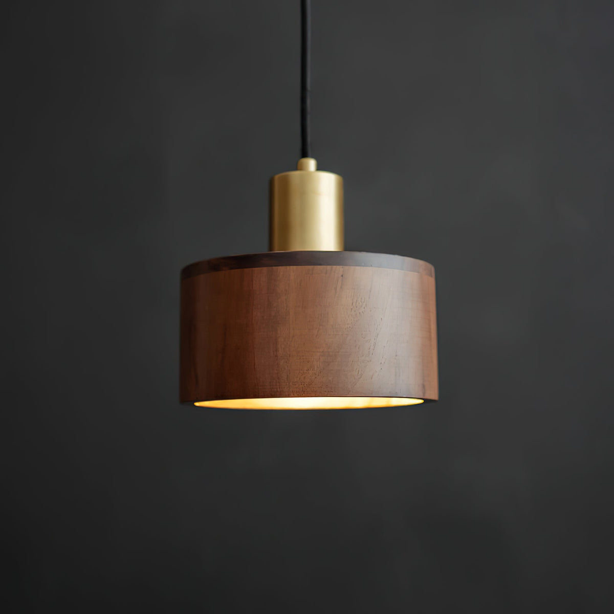 Front view of this Clérachic pendant with a brass cap and a walnut shade, its rim lit softly from below, on a dark backdrop.