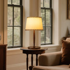 A lit Clérachic table lamp with a beige shade on a round side table beside a tall window and a pale armchair.