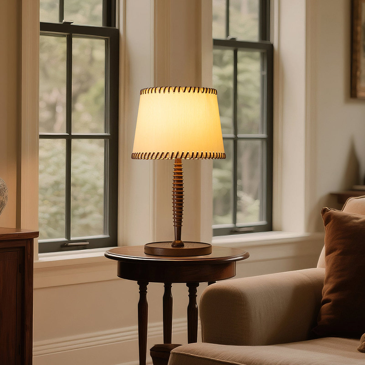 A lit Clérachic table lamp with a beige shade on a round side table beside a tall window and a pale armchair.