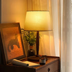 This Clérachic table lamp glows on a wooden bedside chest, next to a framed print, a small plant and a notebook.