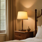 A Clérachic table lamp with a lit beige shade on a wooden bedside table, with a bedpost, curtains and a small lidded jar nearby.