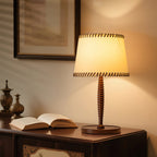 Lit beige shade with dark stitched edges on this Clérachic table lamp, on a wooden sideboard beside an open book.