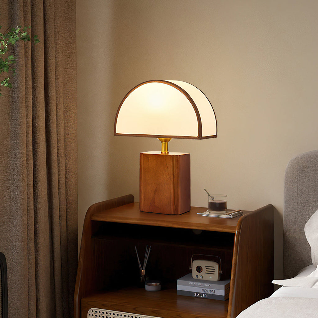 Walnut and white Clérachic table lamp lit on a wooden bedside cabinet, beside a brown curtain and a small radio.