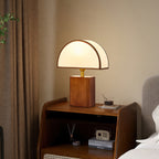 Walnut and white Clérachic table lamp lit on a wooden bedside cabinet, beside a brown curtain and a small radio.