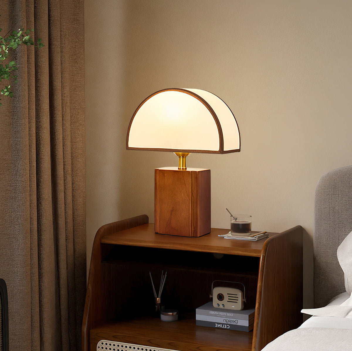 Walnut and white Clérachic table lamp lit on a wooden bedside cabinet, beside a brown curtain and a small radio.