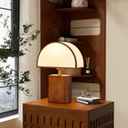 In front of a window with leafy branches, a Clérachic table lamp with a half-round shade stands on a walnut cabinet.
