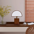 Clérachic table lamp with a half-round white shade on a dark wooden desk, beside a coffee cup, a book and a vase of yellow flowers.