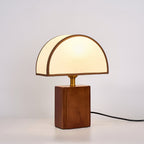 Angled view of a walnut-based Clérachic table lamp with a half-round shade and a black cord trailing out the back.