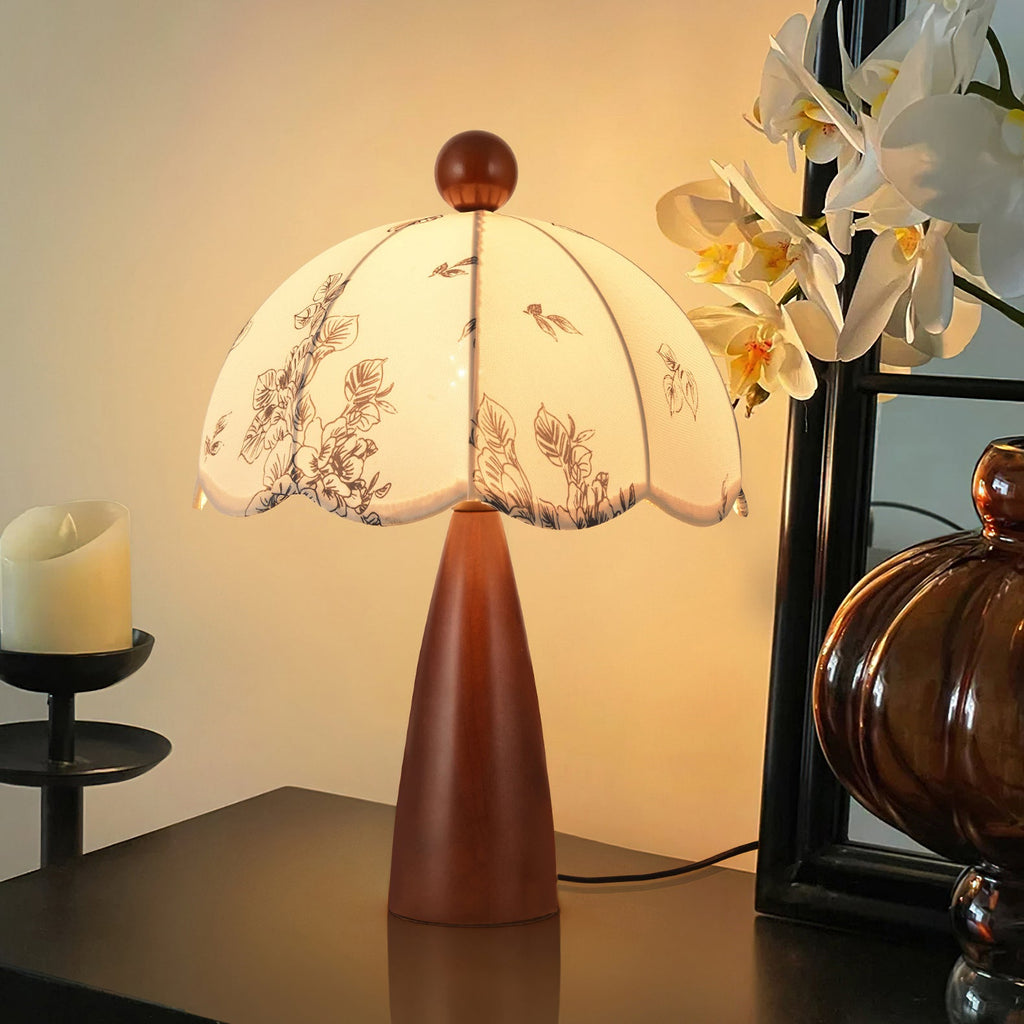 Lit Clérachic table lamp with a tapered wooden base and scalloped shade, on a dark side table beside white flowers.