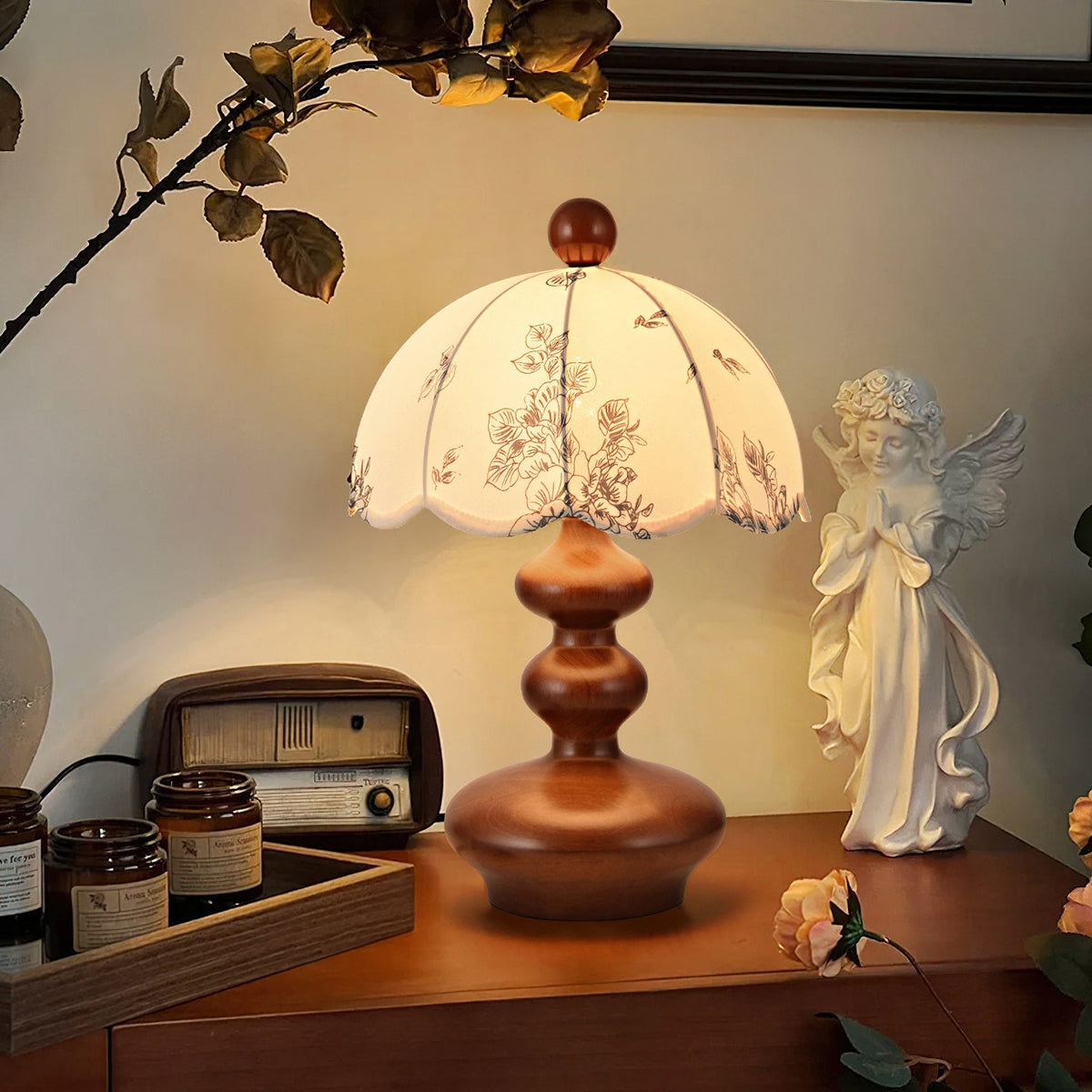 Clérachic table lamp with a rounded, tiered walnut-tone base glowing on a wooden cabinet, with a retro radio and a statuette.