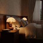 Pair of lit Clérachic lamps with tiered wooden bases on bedside tables, flanking a bed with cream bedding and drawn curtains.