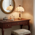 Clérachic lamp with a brass foot and wooden beads on a dark dressing table, repeated in a gilt-framed mirror beside perfume bottles.