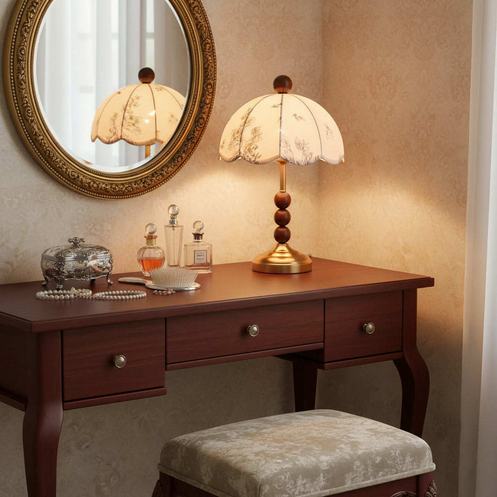 Clérachic lamp with a brass foot and wooden beads on a dark dressing table, repeated in a gilt-framed mirror beside perfume bottles.
