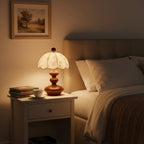 Lit Clérachic lamp with a tiered wooden base on a white bedside table, with a stack of books and a cup by a bed.