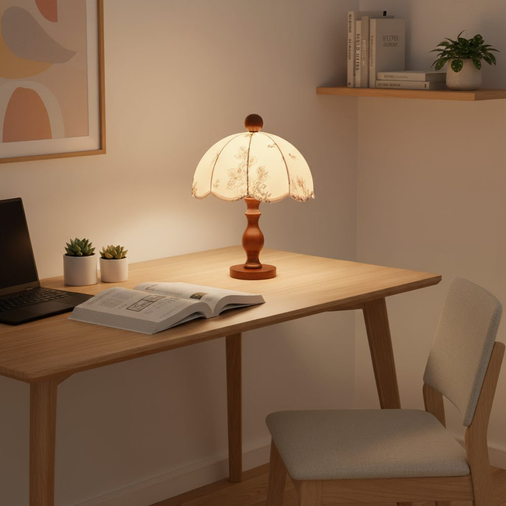 Clérachic lamp with a slim wooden base lit on a light wood desk, with an open book, a laptop and two small plants.