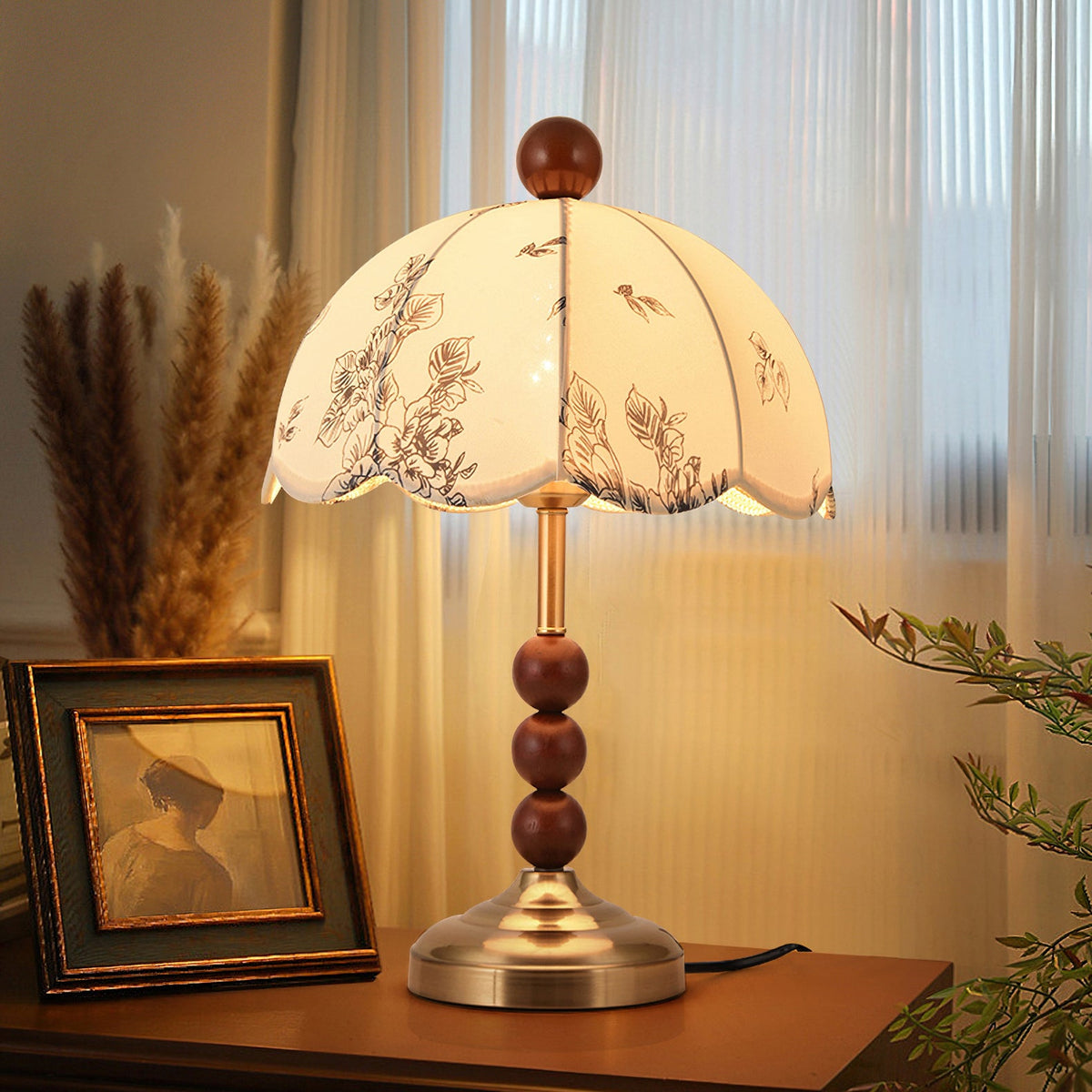 Clérachic lamp with a brass base and wooden bead stem glowing before sheer curtains, beside a framed portrait and dried grasses.