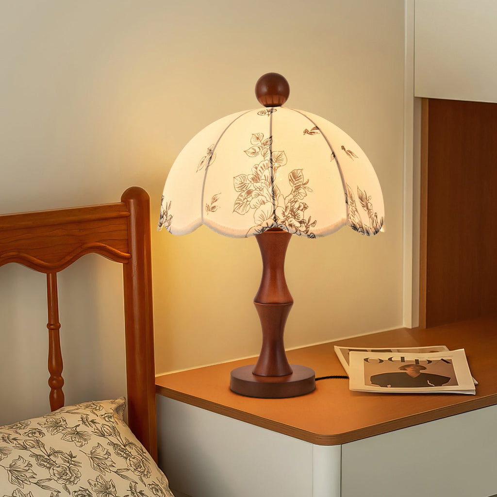 Clérachic lamp with a waisted wooden base lit on a white bedside table, next to a wooden bed frame and a patterned pillow.