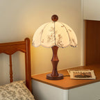 Clérachic lamp with a waisted wooden base lit on a white bedside table, next to a wooden bed frame and a patterned pillow.