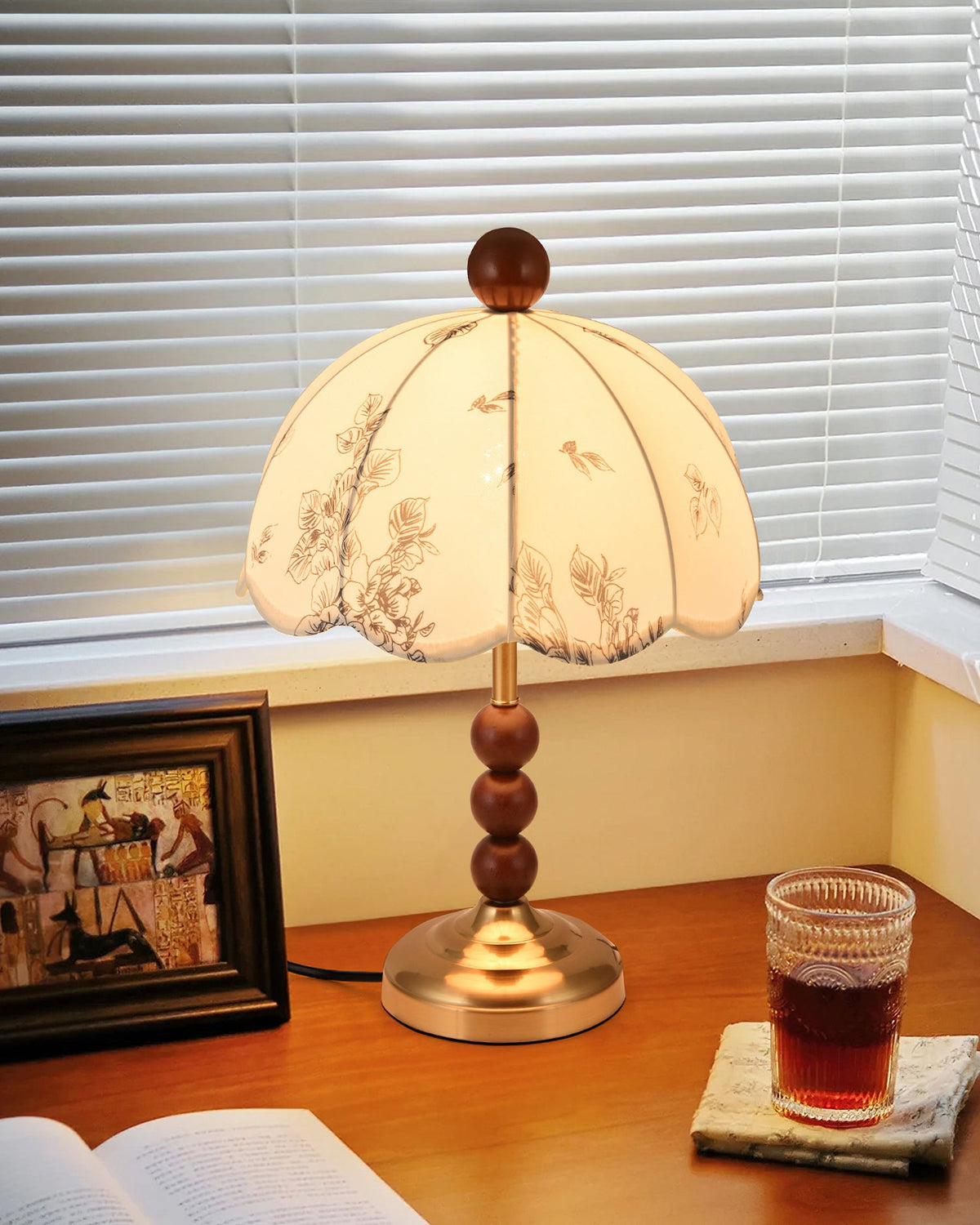 Lit Clérachic lamp with a brass foot on a wooden desk by a window with blinds, with a framed picture and a glass of tea.