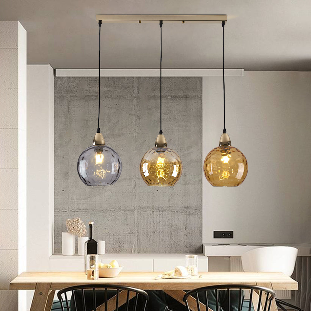 Clérachic 3-globe cluster pendant on its brass bar, lit smoke and amber ripple globes over a wooden dining table.