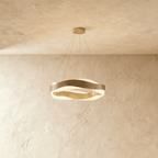 Clérachic round LED chandelier with two wavy gold bands on thin cables, glowing softly against a limewash wall.