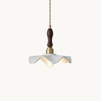 White wavy-edged ceramic shade under a brass socket and a dark carved wooden handle, this Clérachic pendant on a twisted cord against white.