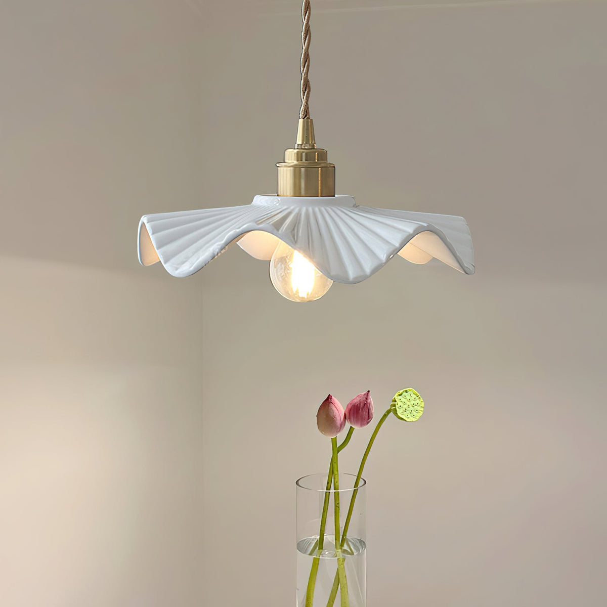 Pleated white ceramic shade with a brass cap on a twisted cord, this Clérachic pendant beside a vase of pink lotus stems.