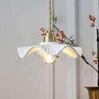 Close, low view of this Clérachic pendant's pleated white shade with its wavy rim, brass cap and a glowing bulb, near berry branches.