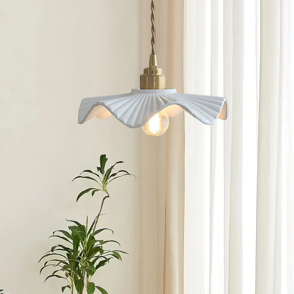 A Clérachic pendant with a rippled white shade beside sheer curtains and a leafy plant, the bulb glowing warm.
