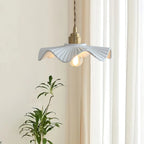 A Clérachic pendant with a rippled white shade beside sheer curtains and a leafy plant, the bulb glowing warm.