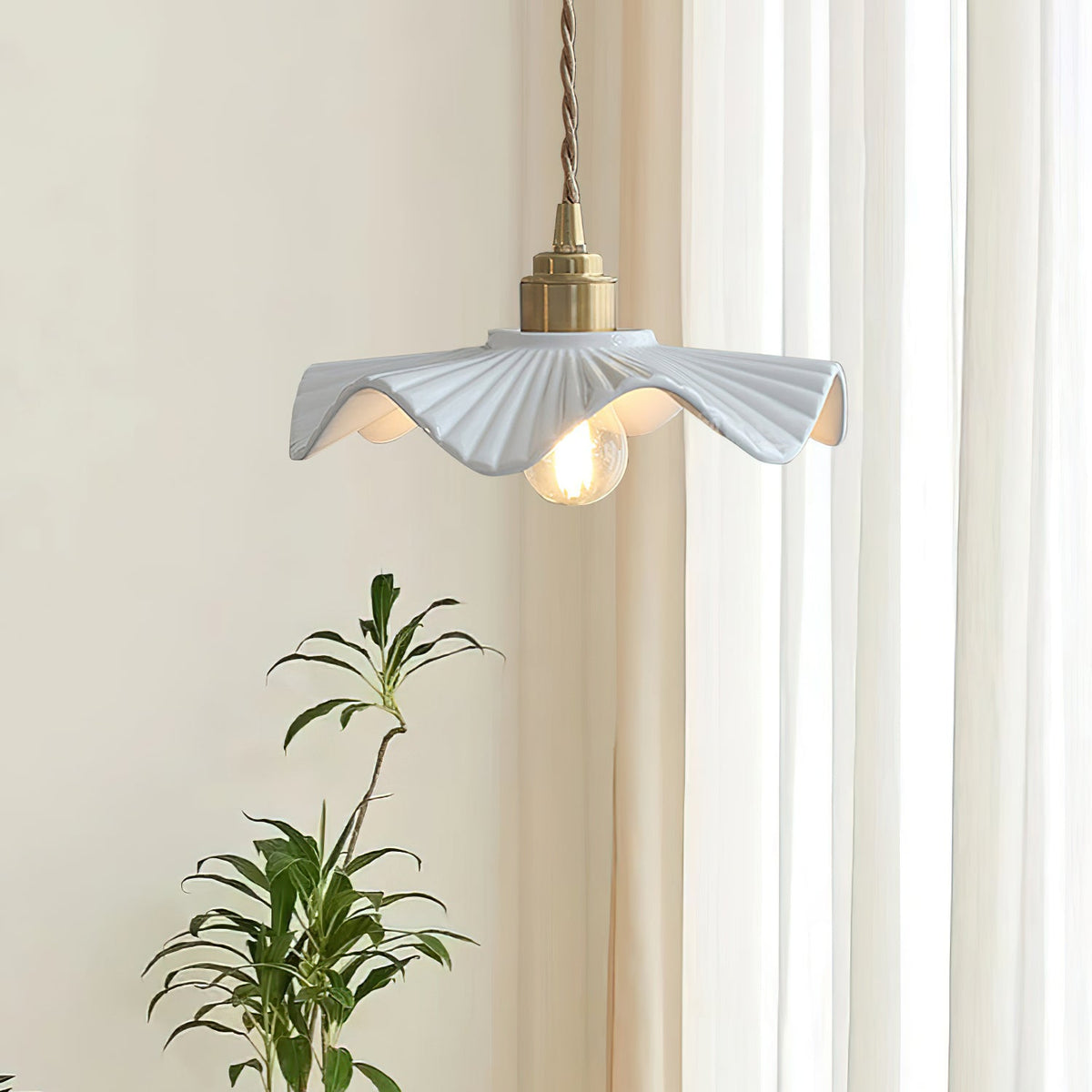 A Clérachic pendant with a rippled white shade beside sheer curtains and a leafy plant, the bulb glowing warm.