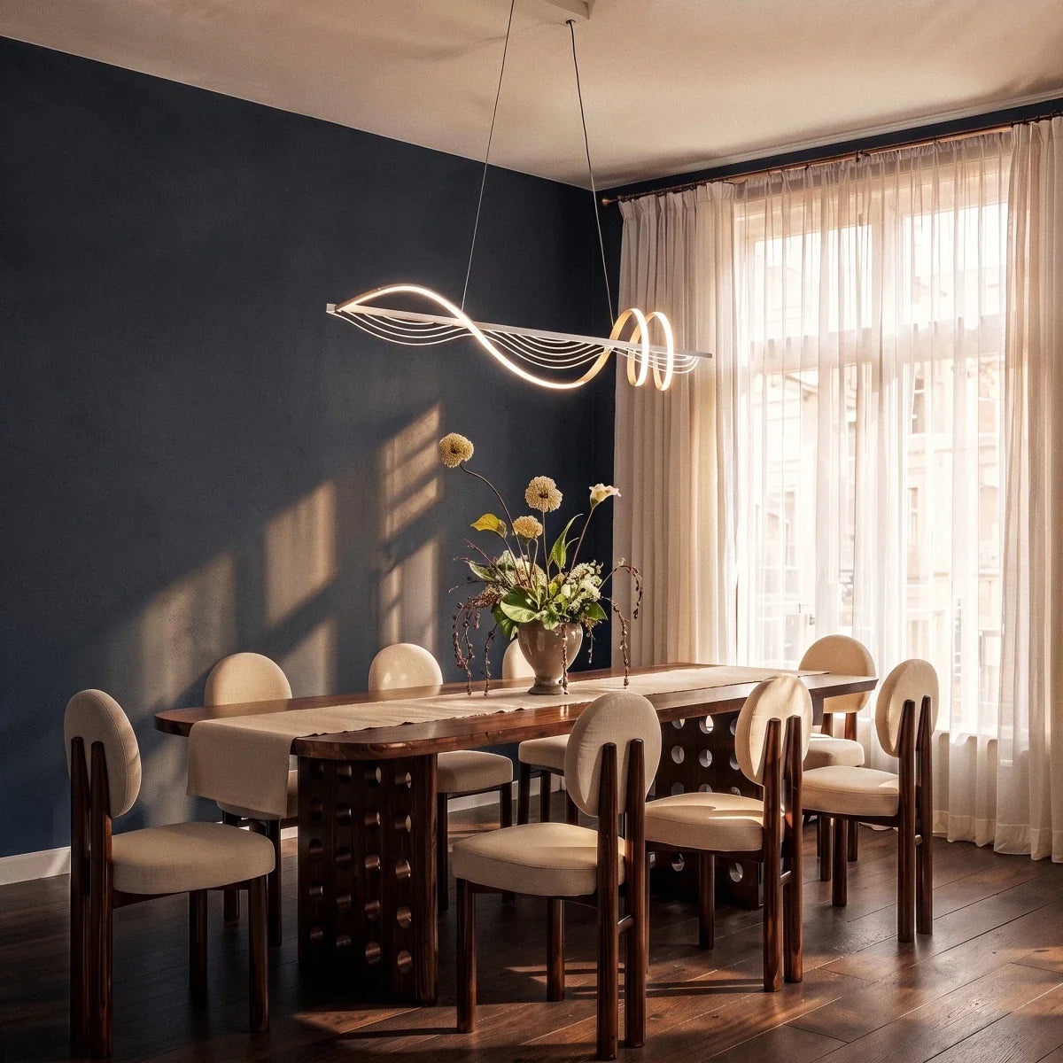 Clérachic white wavy LED pendant over a long walnut dining table with cream chairs, a navy wall and sheer curtains.