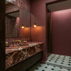 Clérachic pair of wavy wooden pendants glowing warm in a room with burgundy tiled walls, a marble vanity and a mirror.