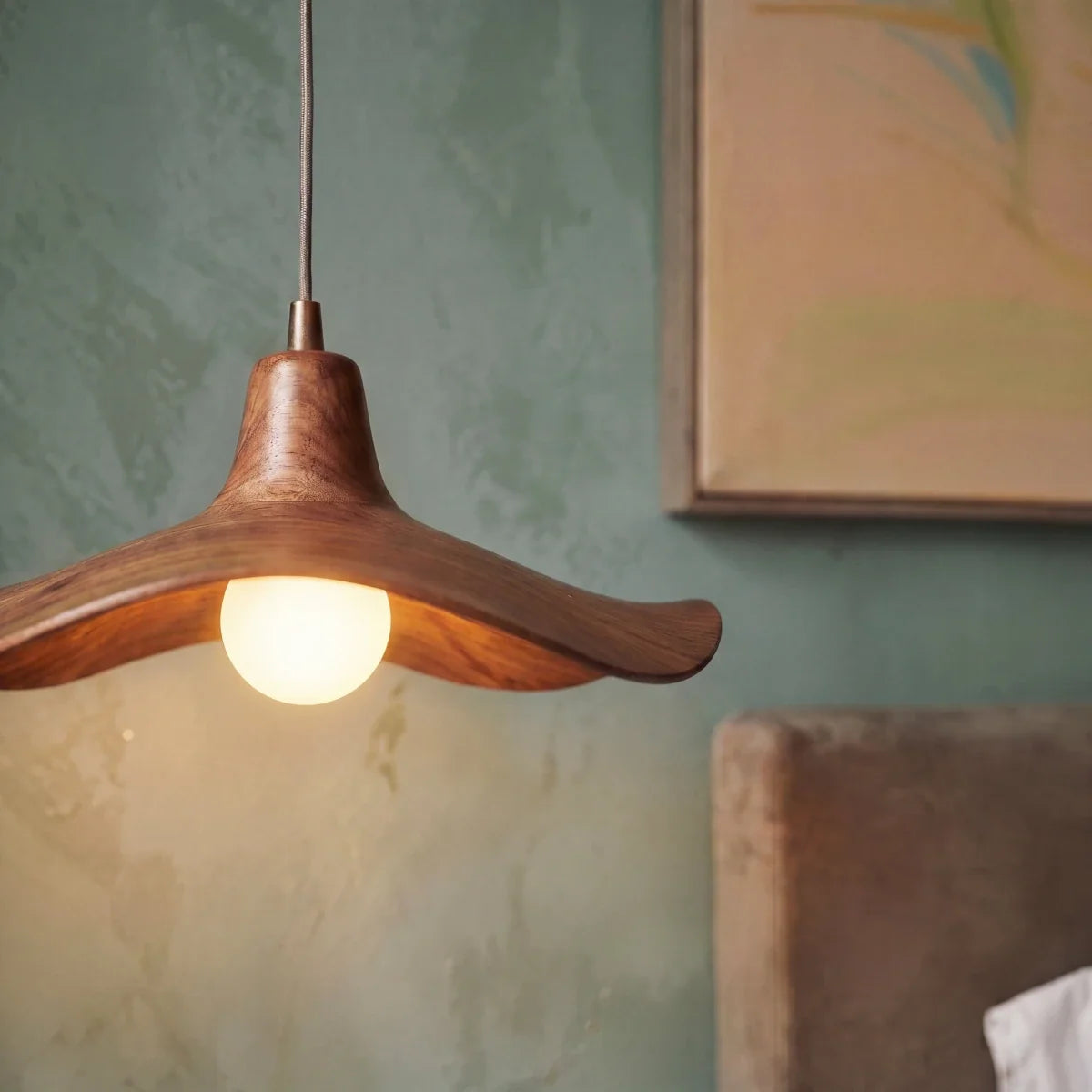Close up of the Clérachic wavy wooden pendant glowing beside a green plaster wall, its frosted bulb lit warm in a bedroom.