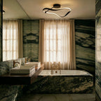 Clérachic black ribbon ceiling light lit above a bathroom with green marble walls, a stone bath, brass taps and linen curtains.