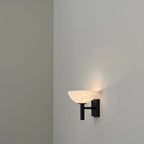 Clérachic Weft sconce switched on in black, throwing a soft upward wash of light across a bare plaster wall.