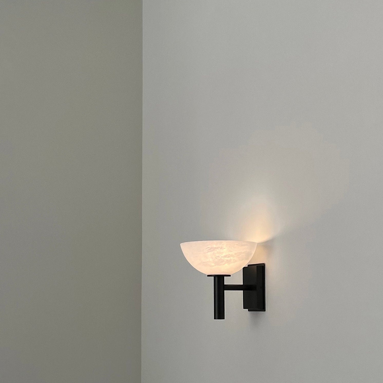 Clérachic Weft sconce switched on in black, throwing a soft upward wash of light across a bare plaster wall.