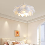 A Clérachic layered-petal pendant lit above a cream bedroom with a tufted headboard, a framed print and a bedside lamp.