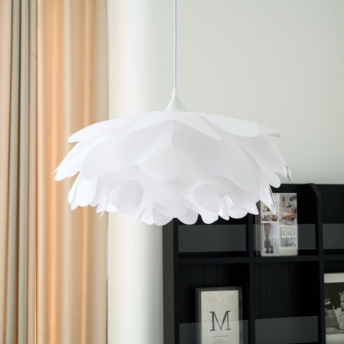 Close view of the overlapping white petals of this Clérachic pendant, unlit, beside a dark shelf and peach curtains.