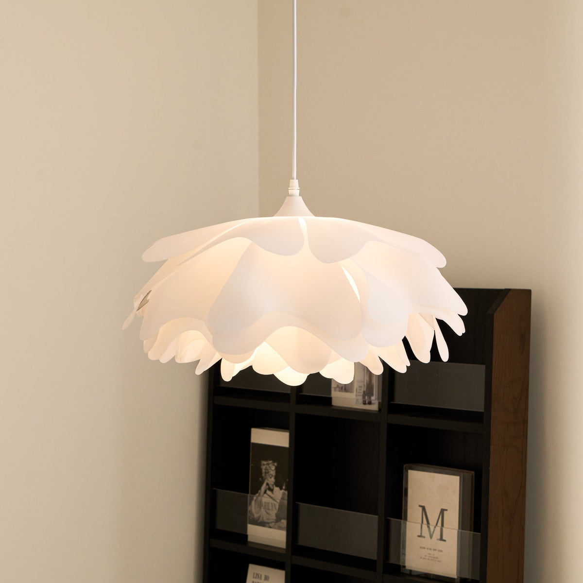 Soft light glowing through the petal layers of a Clérachic pendant, hung in front of a dark shelf with framed prints.