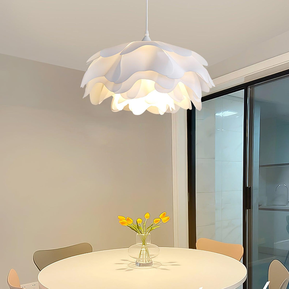 Warm light from a Clérachic pendant with white petals, hung above a round table with yellow tulips beside a sliding glass door.