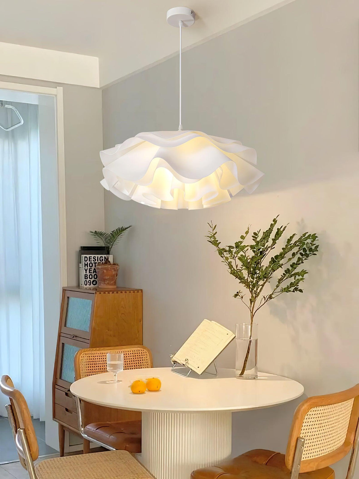 Above a round white table with cane chairs, this Clérachic pendant glows beside a wooden cabinet and a leafy branch in a vase.