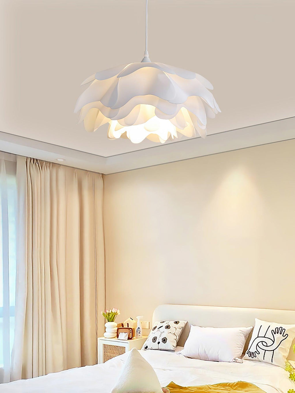 A Clérachic pendant glowing above a bedside table with patterned cushions, beside beige curtains in a warm bedroom.