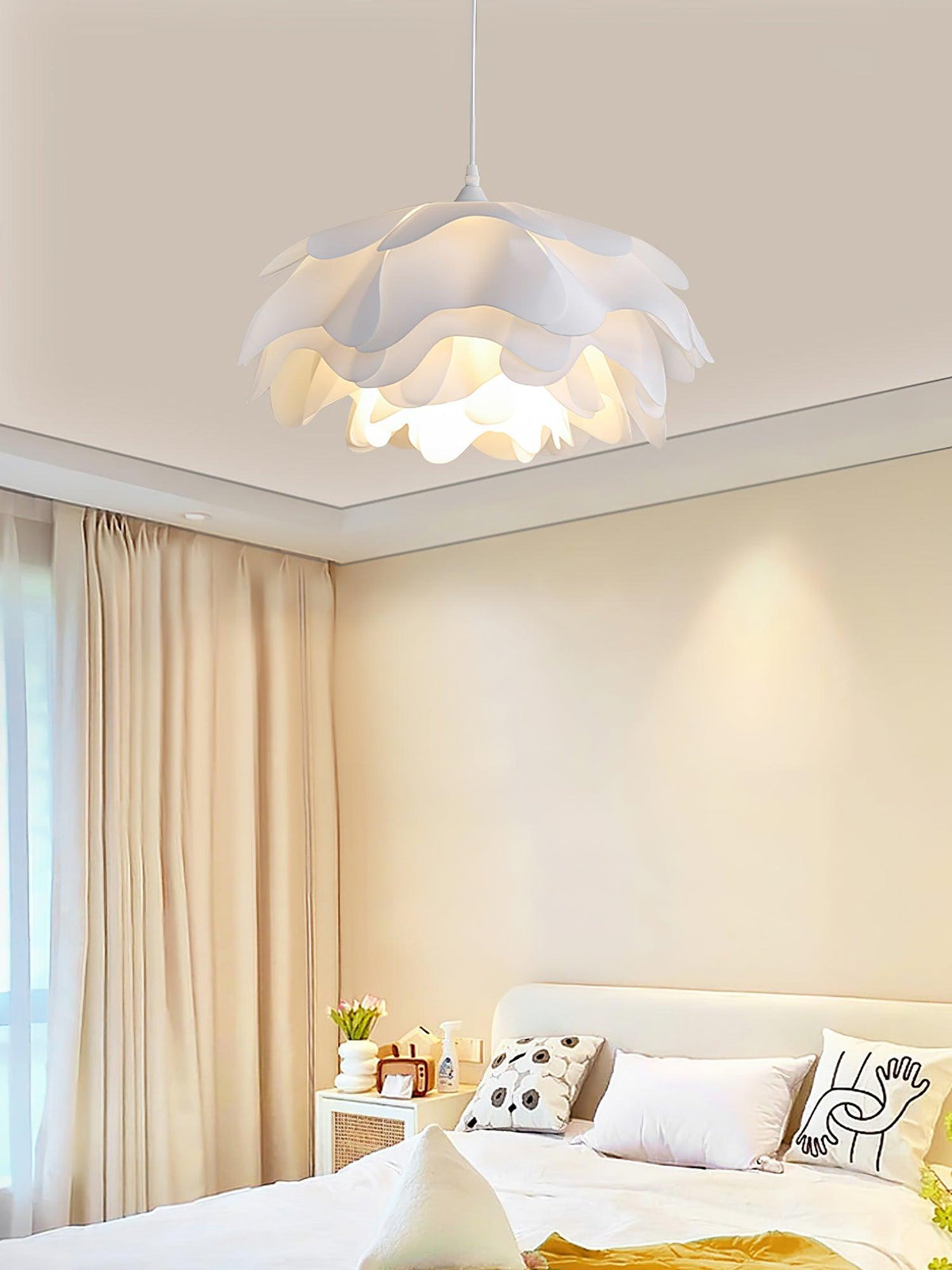 A Clérachic pendant glowing above a bedside table with patterned cushions, beside beige curtains in a warm bedroom.