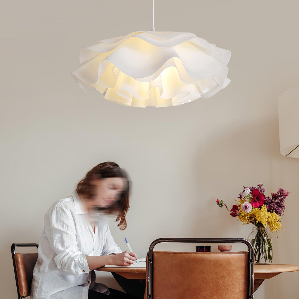 A woman writing at a wooden table below this Clérachic pendant, whose ruffled white petals glow against a beige wall.
