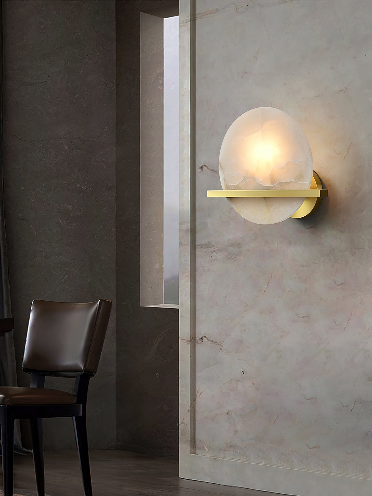 The Clérachic White Alabaster Wall Sconce lit on a stone-clad dining wall, casting a soft halo beside a dark leather chair.