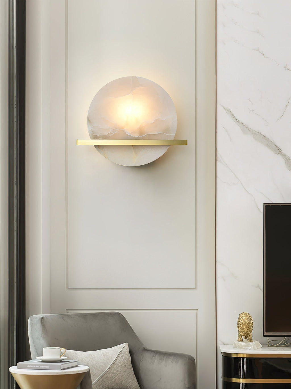 Clérachic White Alabaster Wall Sconce glowing on a cream panelled wall above a grey velvet armchair in a bright sitting room.