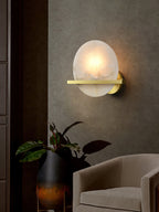 Clérachic White Alabaster Wall Sconce glowing on a warm grey wall in a dim lounge, beside a potted plant and a cream armchair.