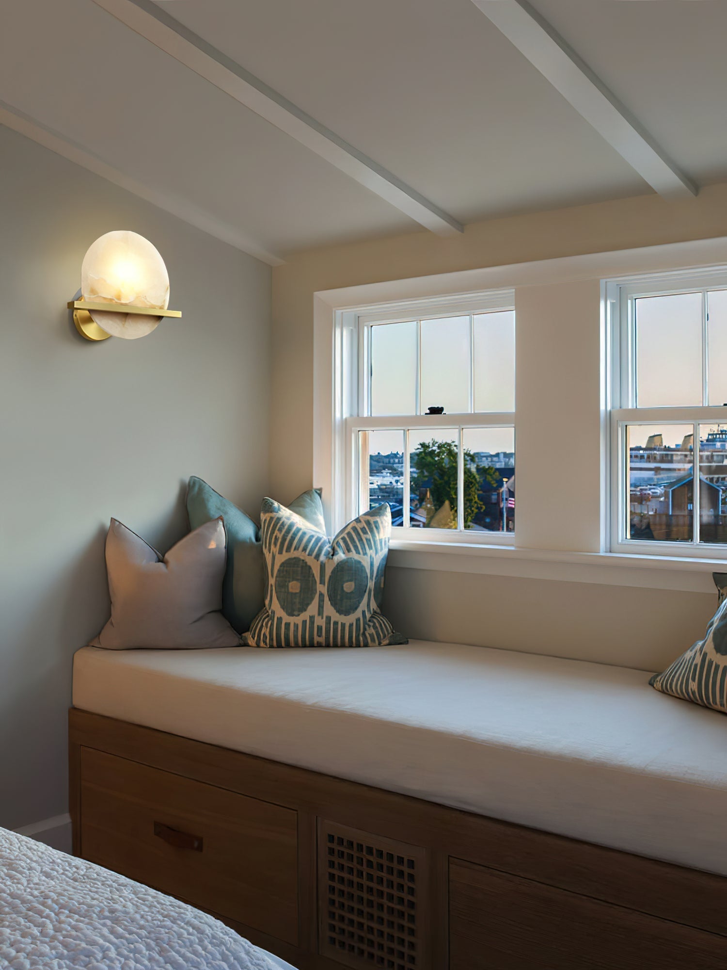 Clérachic White Alabaster Wall Sconce lit in a beamed bedroom, throwing a soft pool of light over the cushioned window seat.