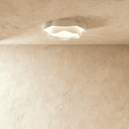Clérachic low-profile white ceiling light with ruffled edges, glowing warm on a pale textured ceiling above a limewash wall.